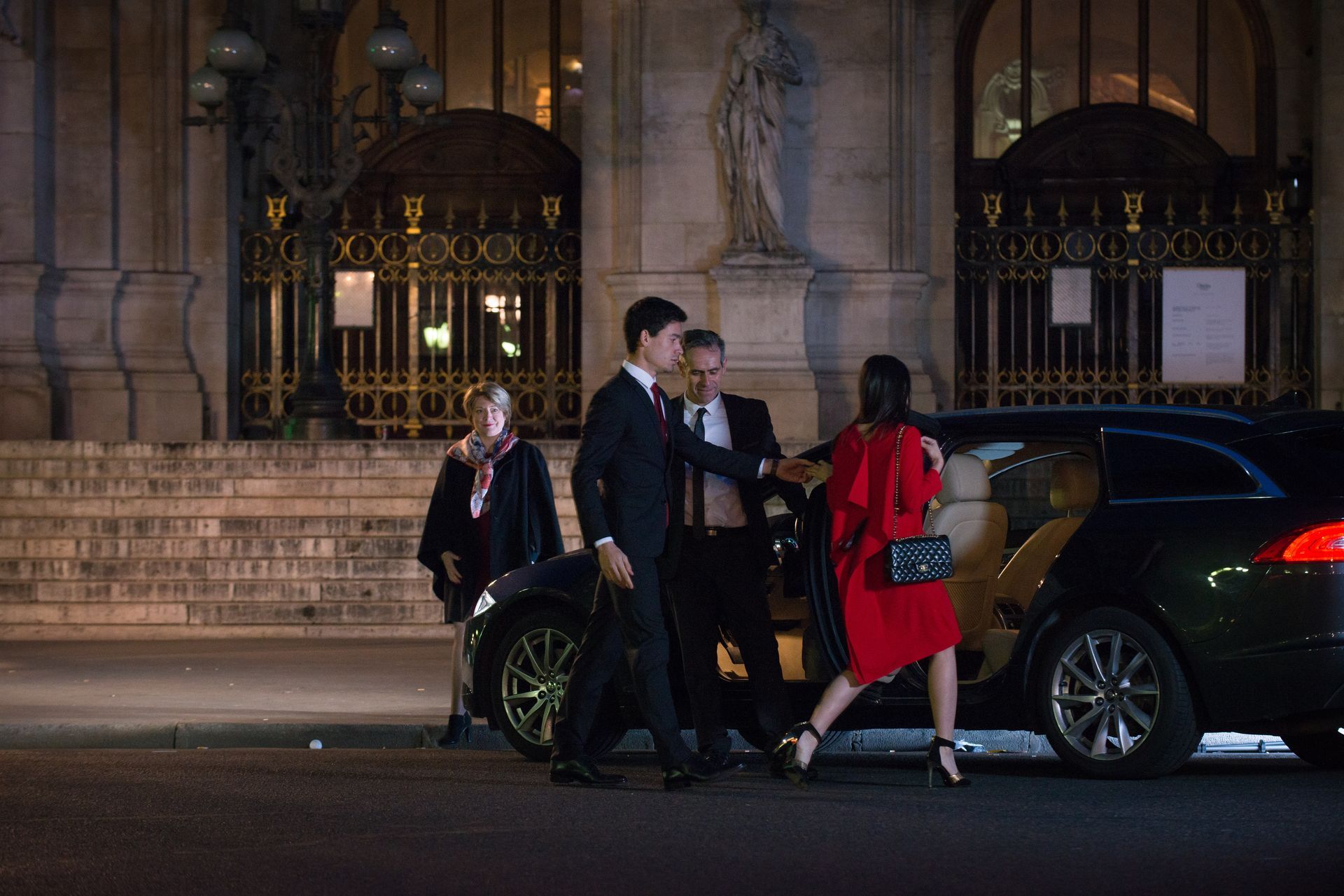 People exiting a car outside a building at night. Woman in red coat gets help, others in suits.