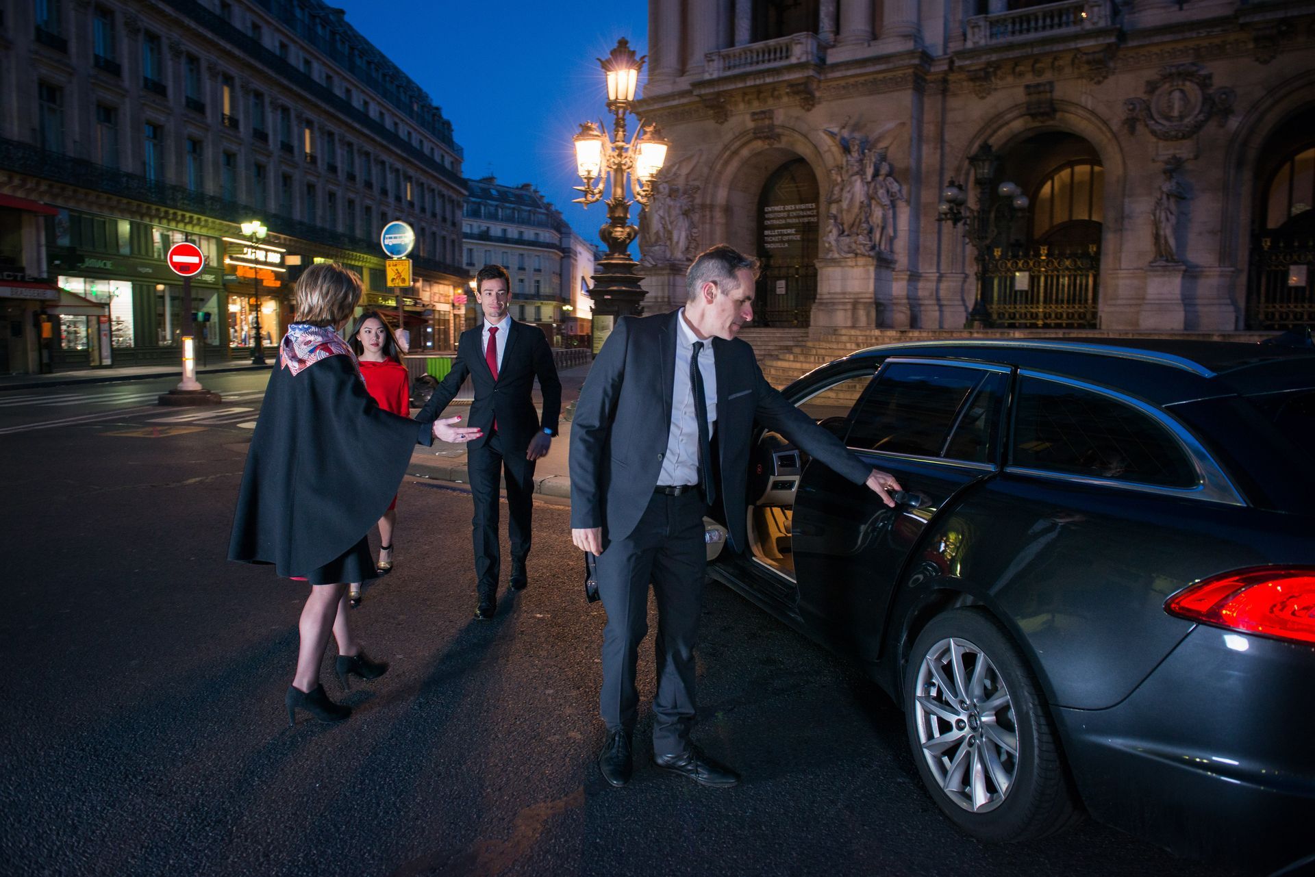 Man in suit opens car door for person in cape in front of a building at night.