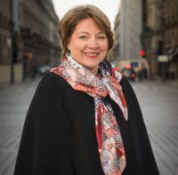 Woman in black coat and scarf smiling in a city street.
