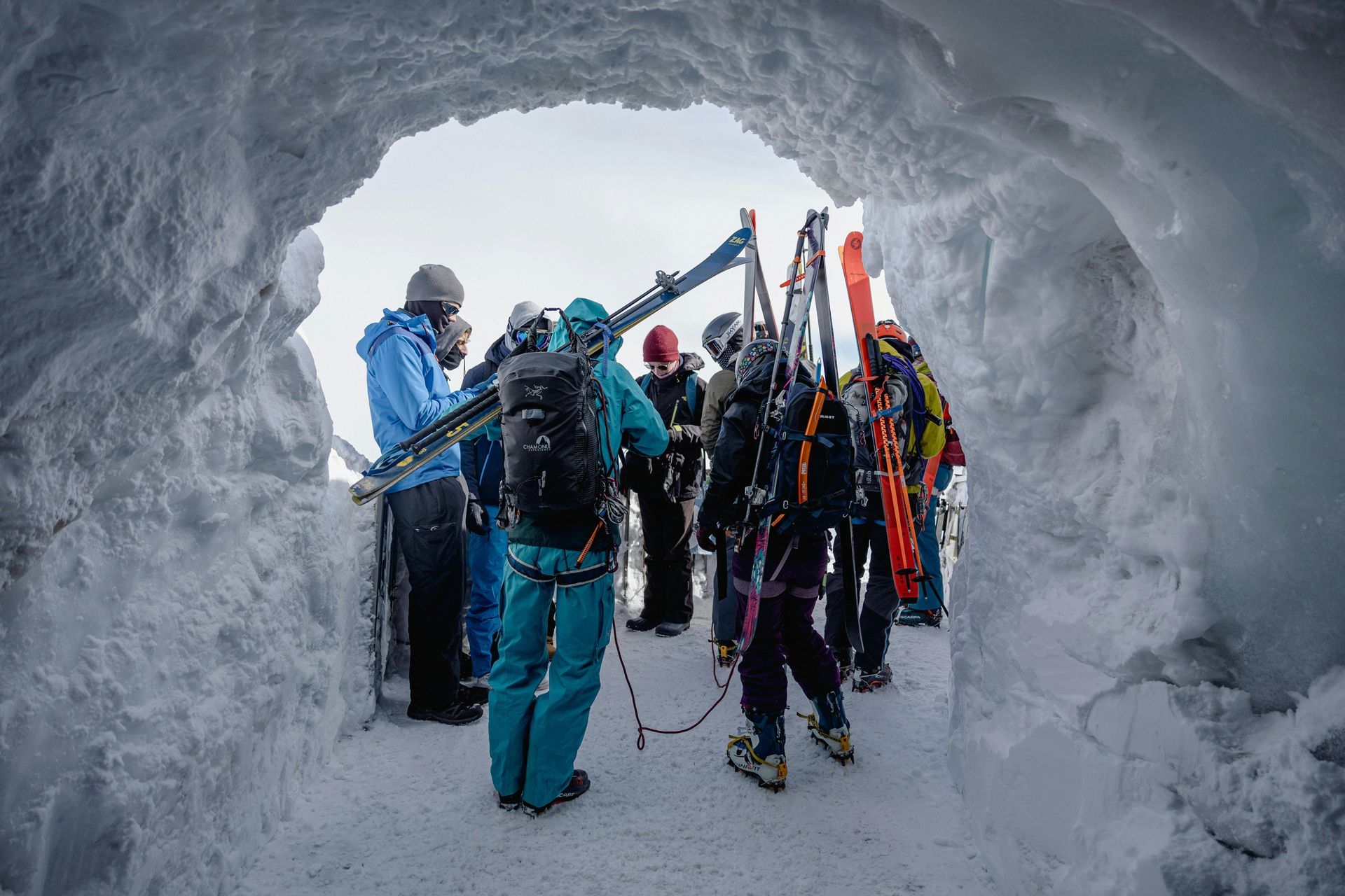 People with skis exit a snowy tunnel into a cloudy outdoor scene.