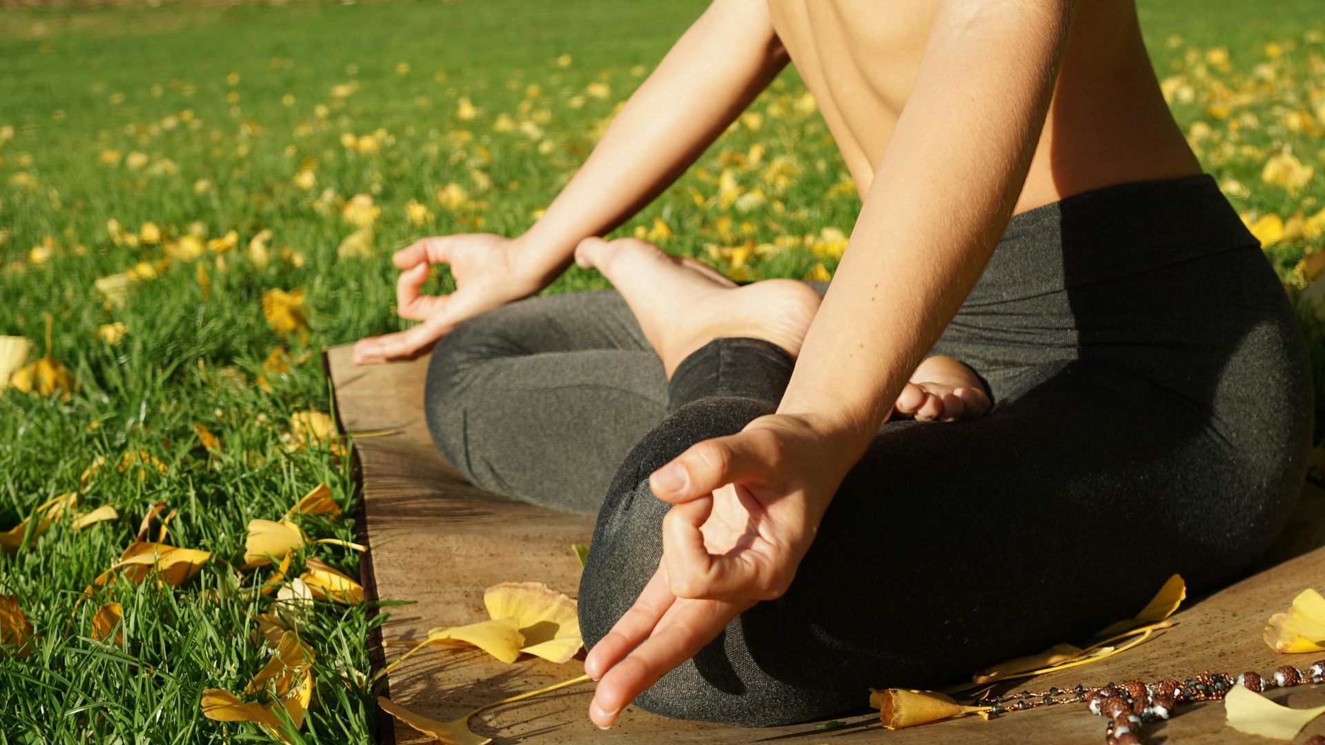 Person meditating outdoors, hands in mudra, seated on a mat, surrounded by yellow leaves.