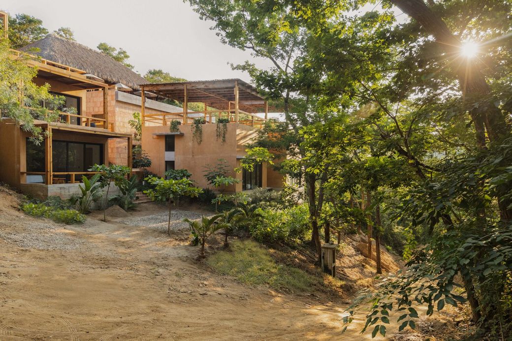 Rustic houses on a hillside surrounded by trees, with a dirt path in the foreground and sunlight filtering through foliage