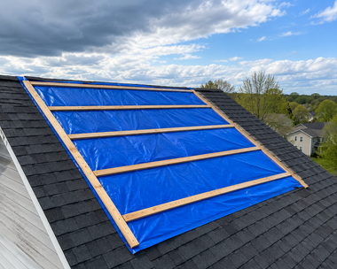 Blue tarp secured over roof section with wooden battens under a cloudy sky