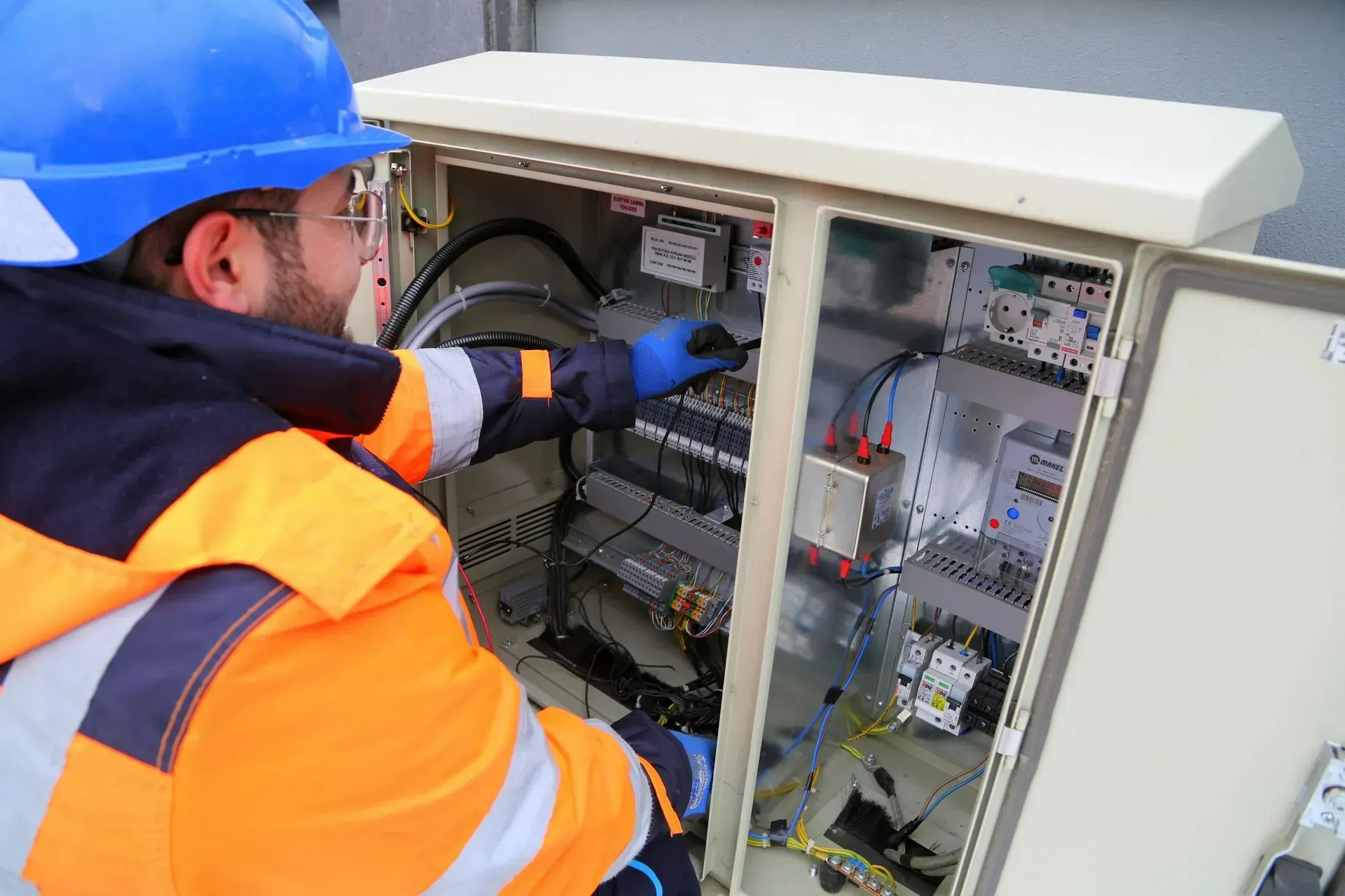 Technician in a blue hard hat and high-visibility jacket inspecting electrical wiring inside an open utility cabinet.