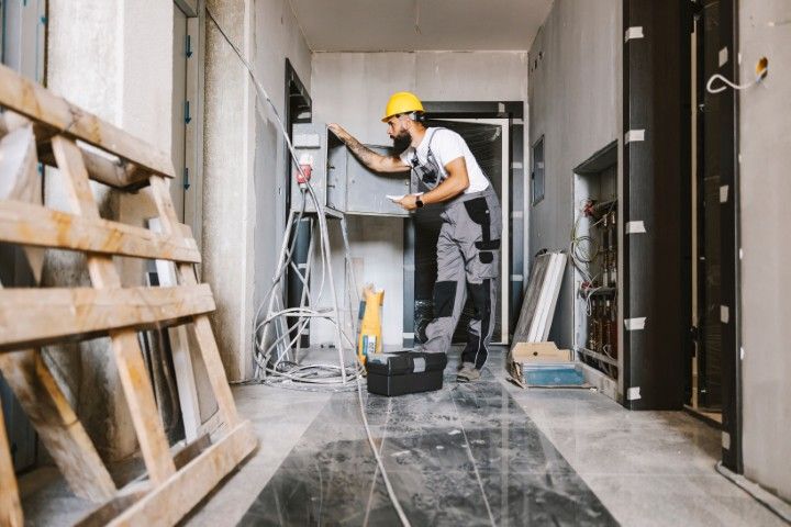 An electrician in a hard hat and grey overalls works on a wall-mounted electrical panel in a hallway under construction.