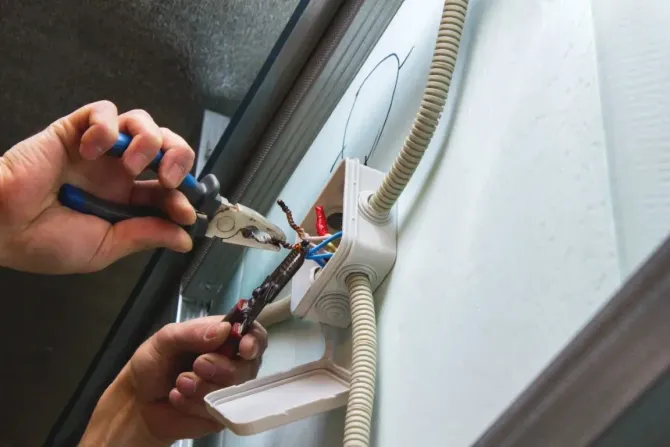 Hands using pliers to work on exposed electrical wiring inside a wall-mounted junction box.