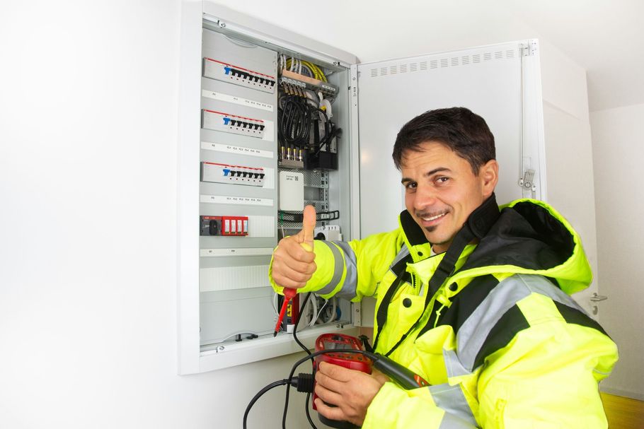 A technician in a high-visibility yellow jacket giving a thumbs-up while working on an open electrical circuit breaker box.