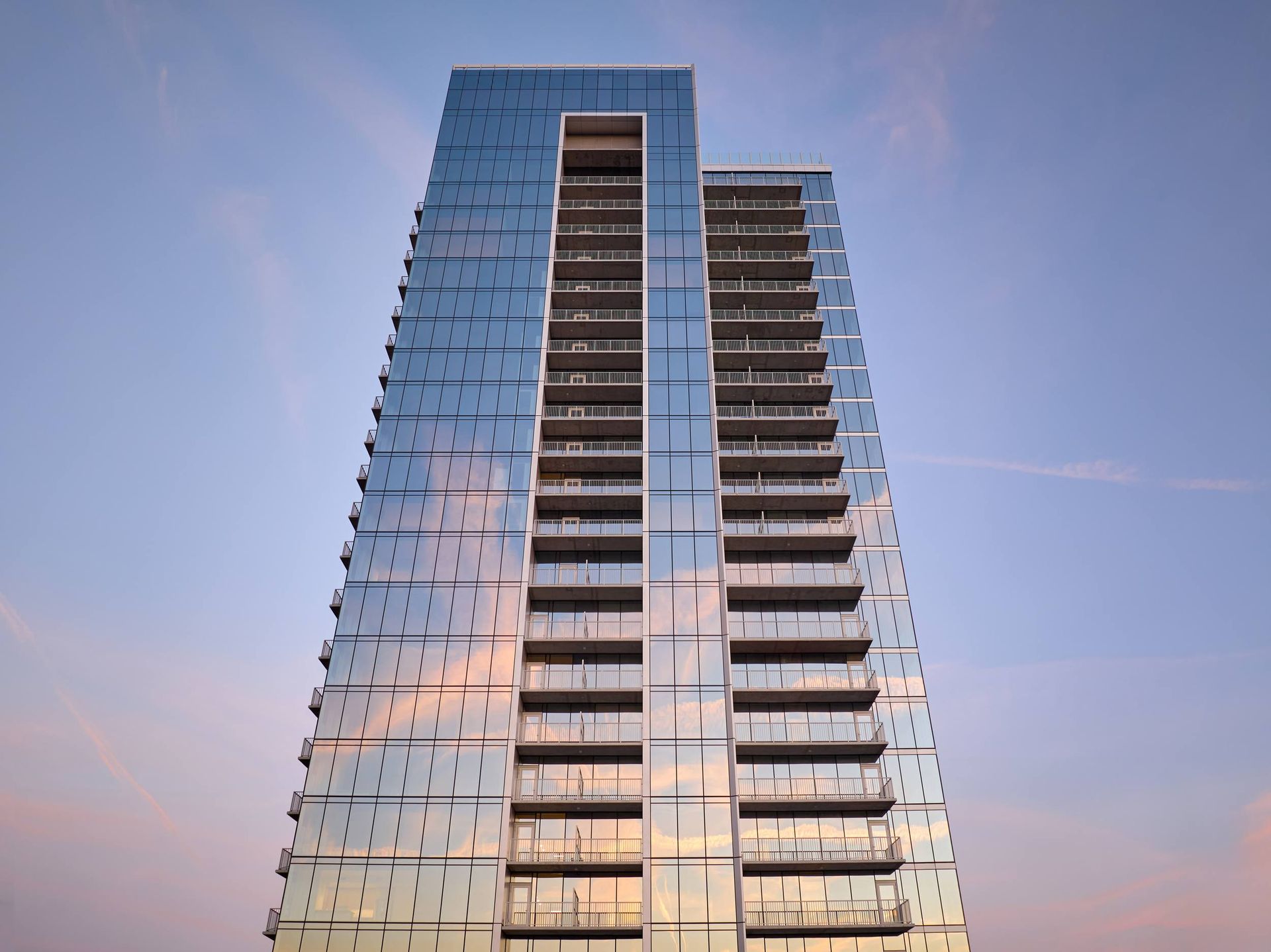 Tall, modern glass skyscraper against a blue and pink sunset sky. Many balconies visible.