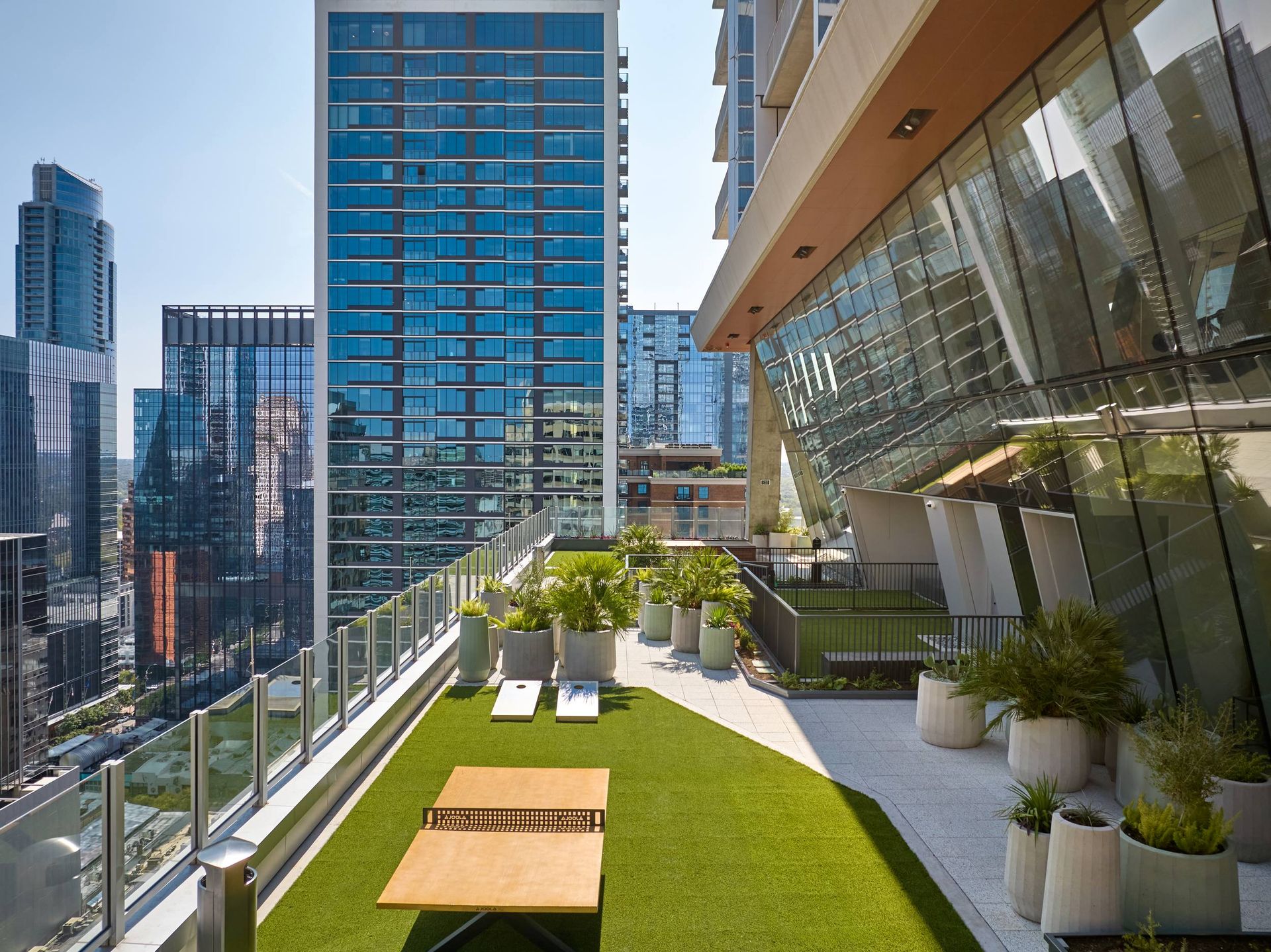 Rooftop terrace with artificial grass, plants, table, and cornhole boards overlooking cityscape of modern buildings.