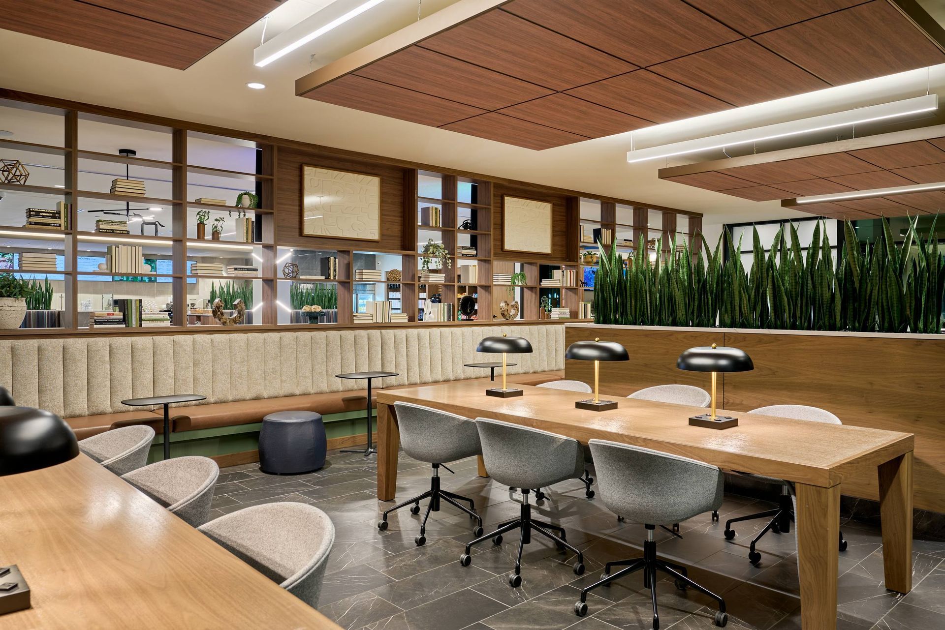 Workspace with desks, chairs, lamps, and a shelf filled with items. Brown wood ceiling and paneling, green plants.