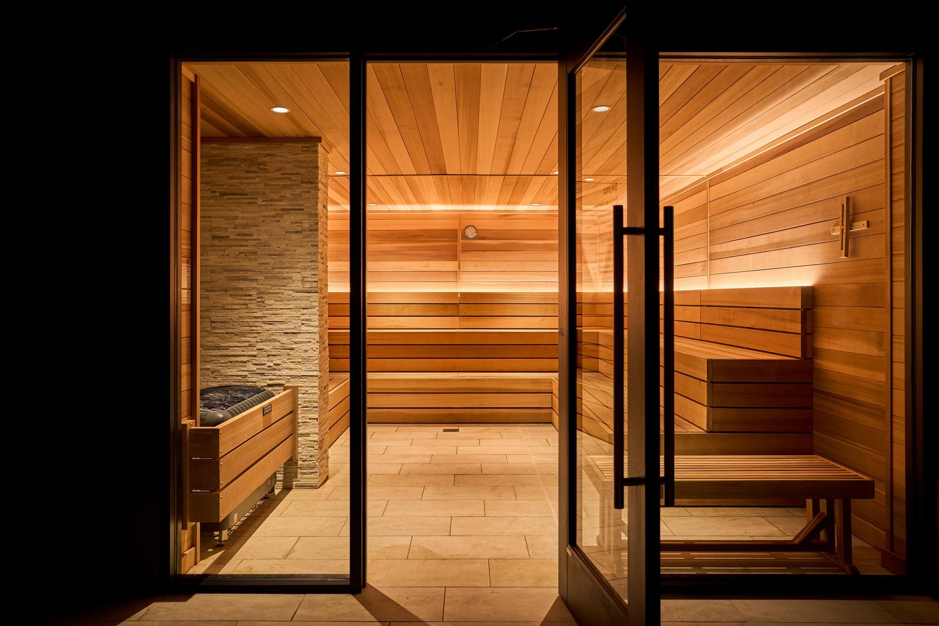 Wooden sauna interior with open glass door, warm lighting, benches, and stone wall.