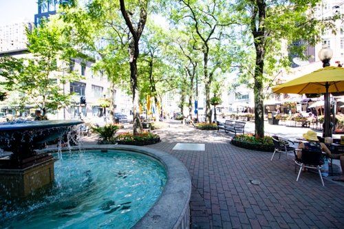 A fountain in a park with people sitting at tables under yellow umbrellas.