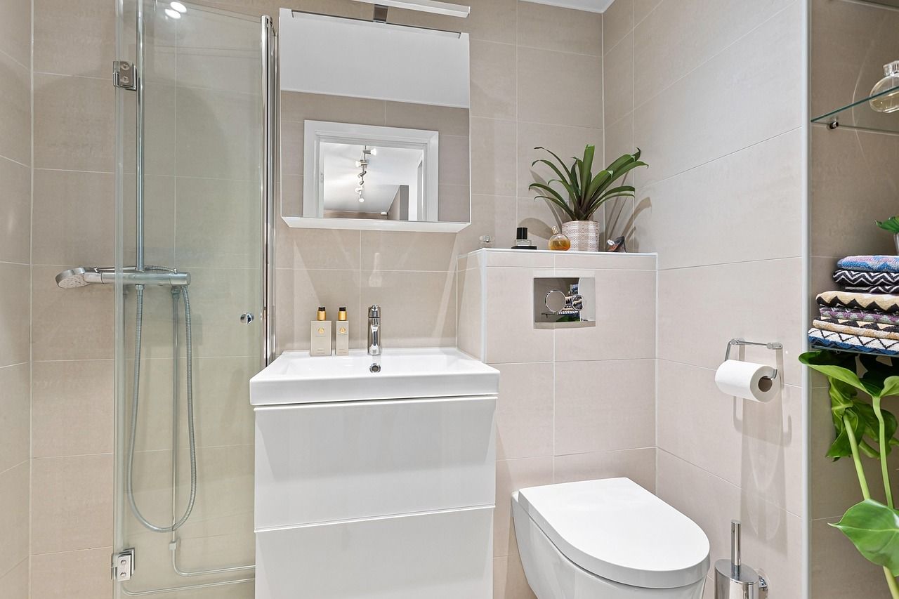 Small, modern bathroom. White sink, toilet, and shower. Beige tile walls. Plant on shelf.