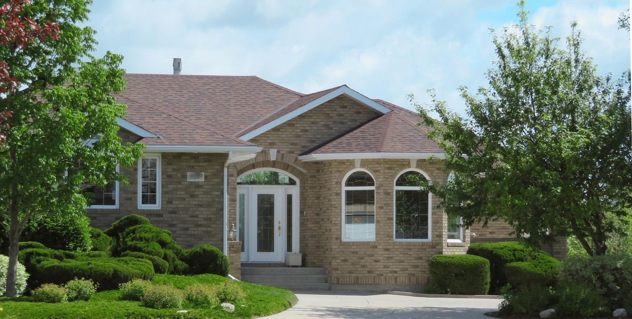 Tan brick house with brown roof, white arched windows, and front door. Green bushes and trees in front.