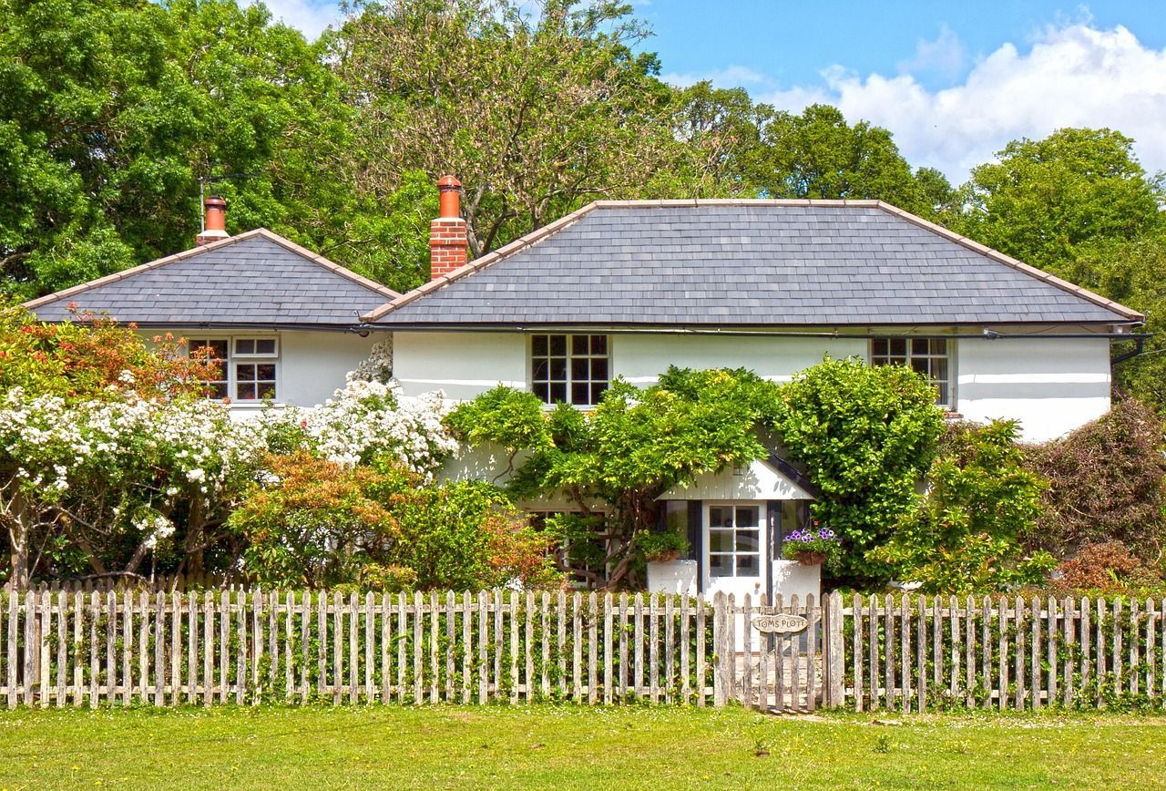 White cottage behind a picket fence, surrounded by lush greenery under a bright blue sky.