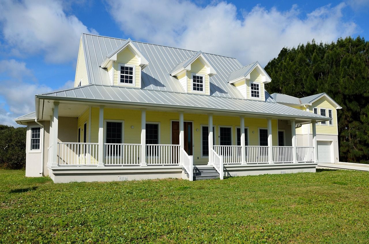 Yellow house with white porch and railing, gray roof, green lawn under blue sky.