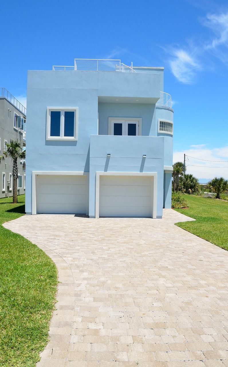 Two-story light blue beach house with two garage doors and a curved paved driveway, blue sky.