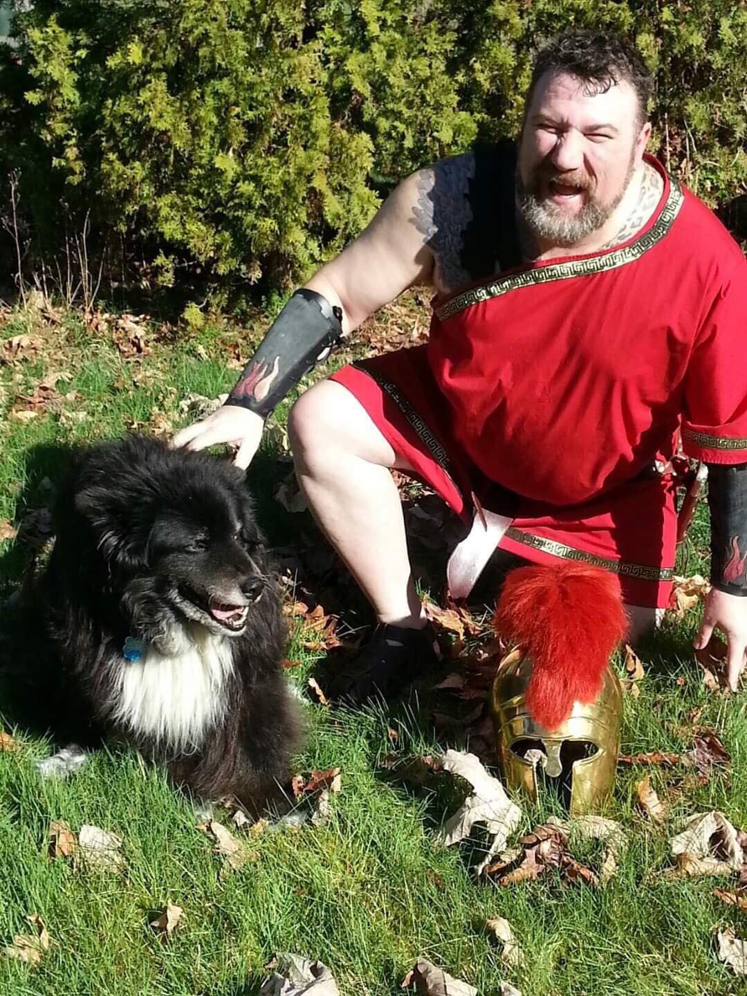 A man in a red shirt is kneeling down next to a black and white dog, representing Ares.
