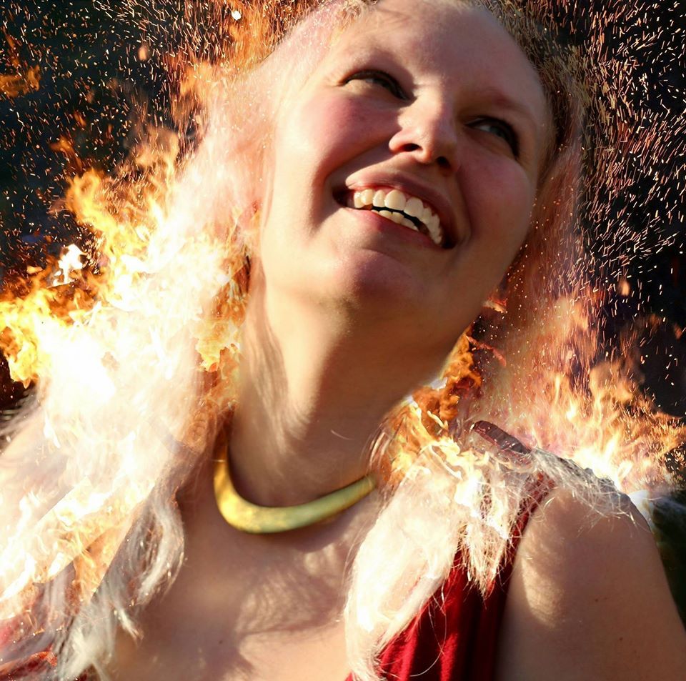 A woman with flames coming out of her hair is smiling,  representing Hestia.