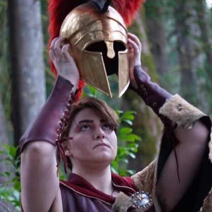 A woman in a spartan costume is putting on a helmet, representing Ares.