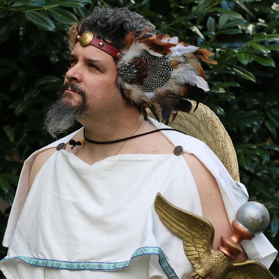A man with a beard is wearing a feathered headdress, representing Hermes.