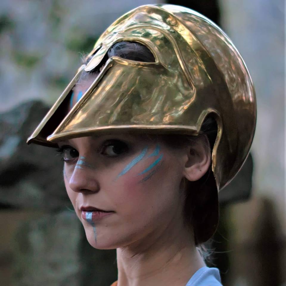 A woman wearing a brass helmet with blue paint on her face, representing Athena.