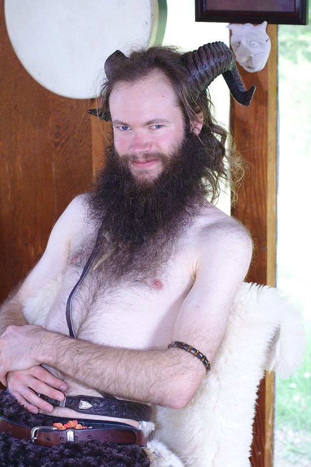 A shirtless man with a beard and horns is sitting in a chair,  representing Pan.