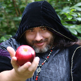 A man in a black hood is holding a red apple, representing Hades.