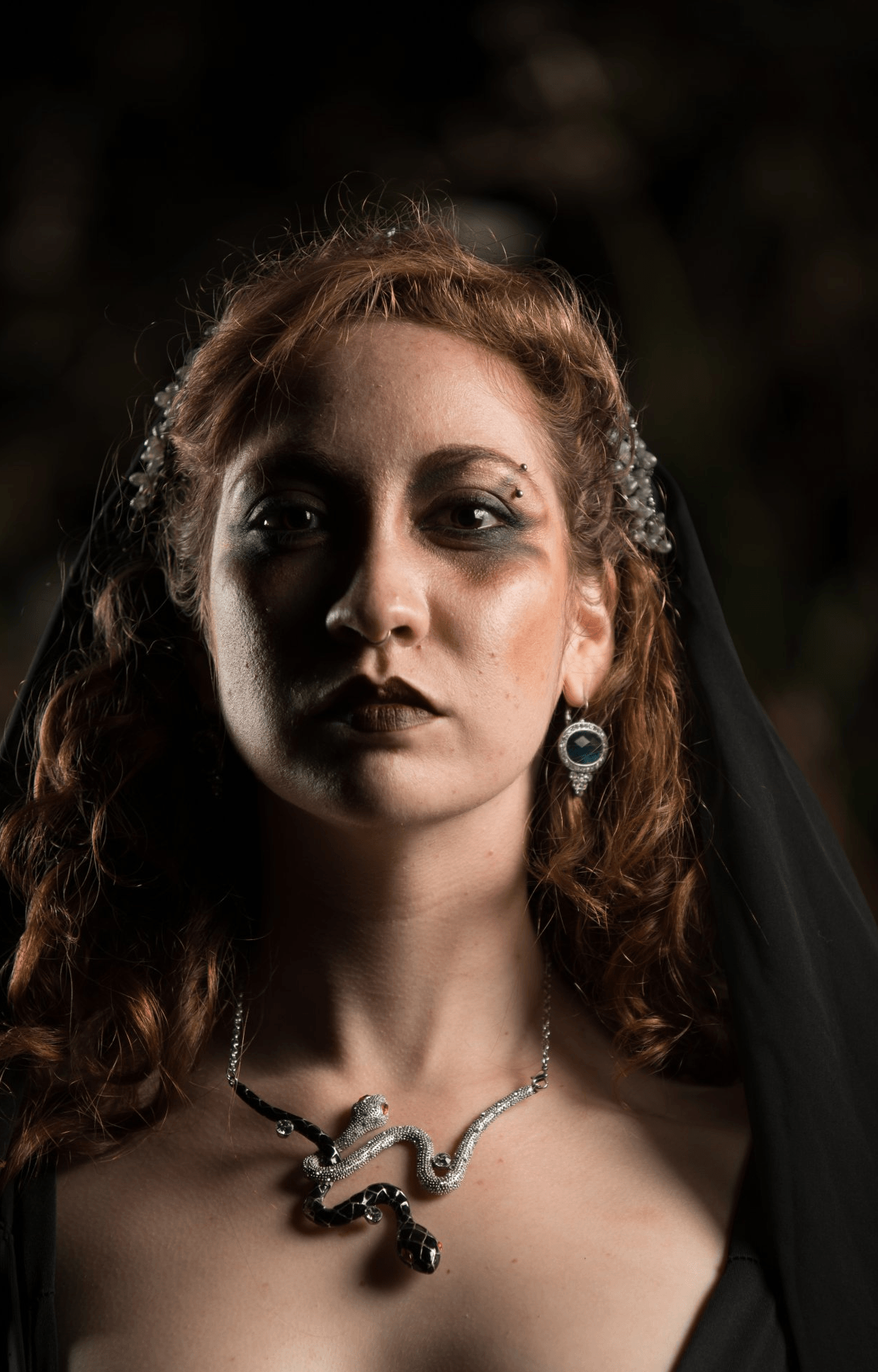 A woman wearing a black veil and a necklace is looking at the camera, representing Hekate.