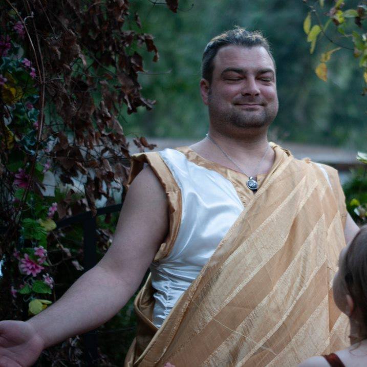 A man in a white and gold robe is smiling, representing Apollo.