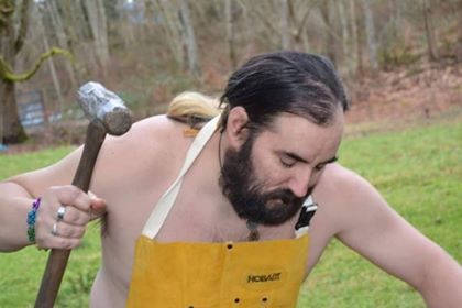 A shirtless man with a beard is holding a hammer,  representing Hephaestus.
