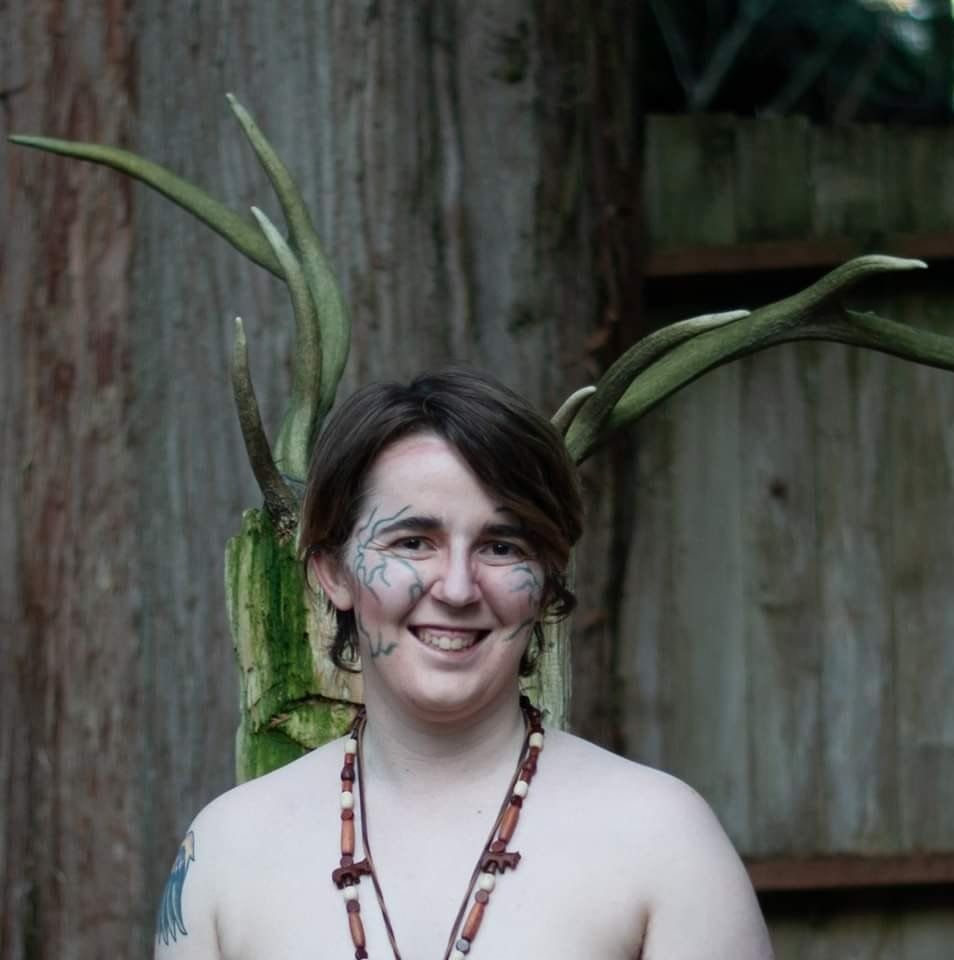 A woman without a shirt has a tattoo on her face,  representing Pan.
