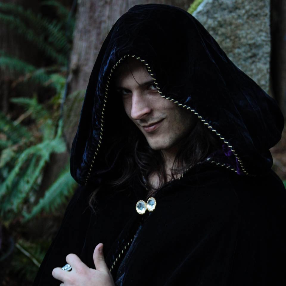 A man wearing a black cape with a hood and a ring on his finger,  representing Hades.