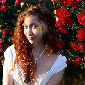 A woman with a flower painted on her face is standing in front of a bush of red roses, representing Aphrodite.