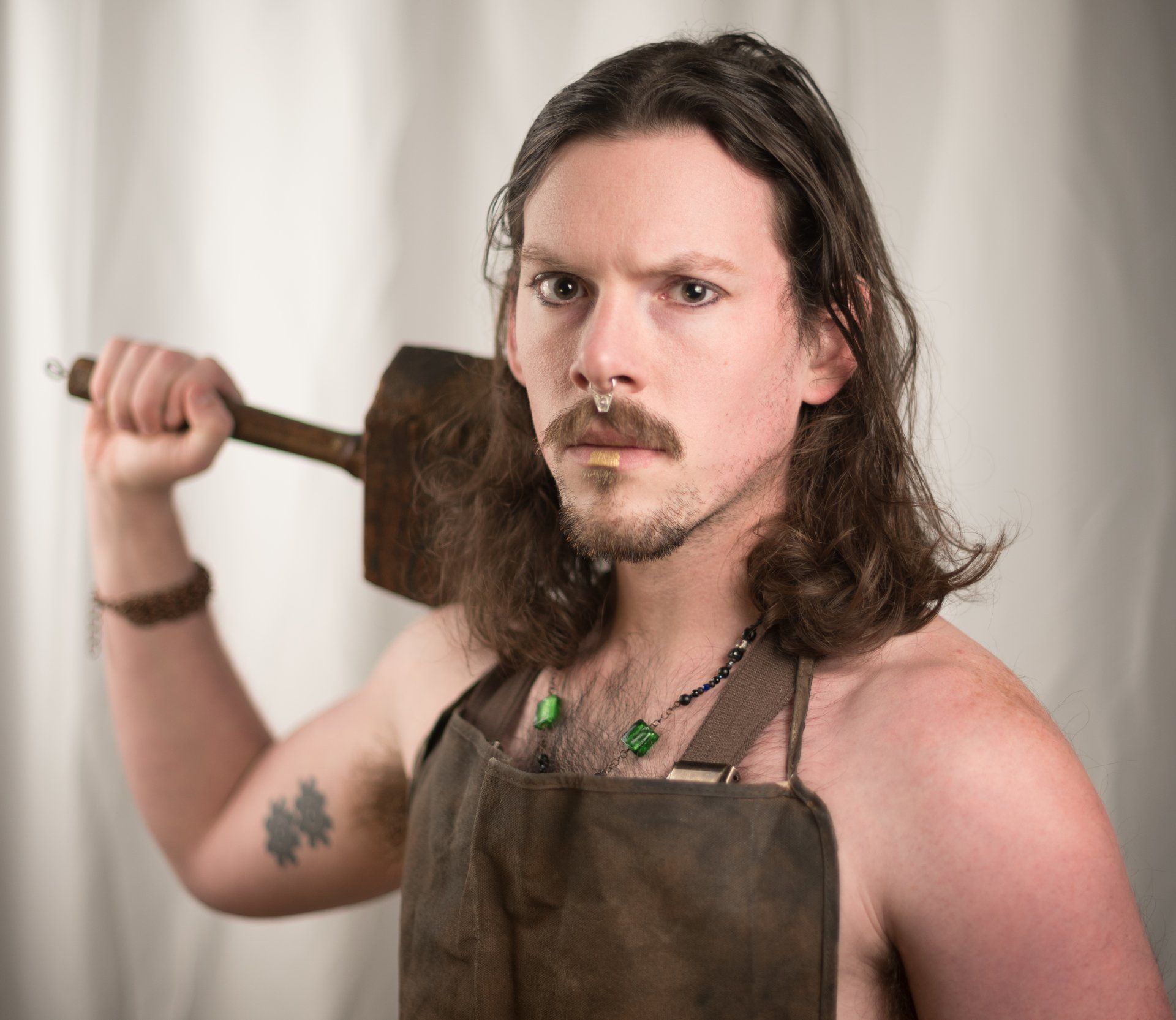 A shirtless man in an apron is holding a large hammer over his shoulder,  representing Hephaestus.