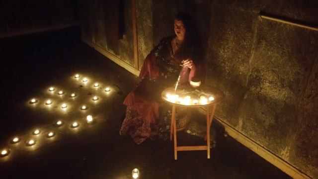 A woman sitting at a table with candles on the floor in the dark representing Hekate.