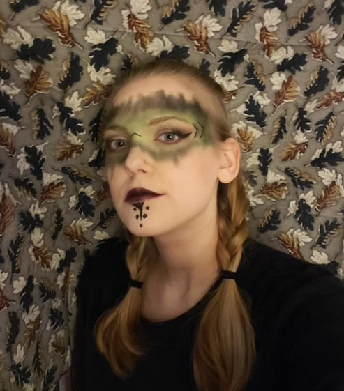 A woman with makeup on her face is standing in front of a wall with leaves on it, representing Artemis.
