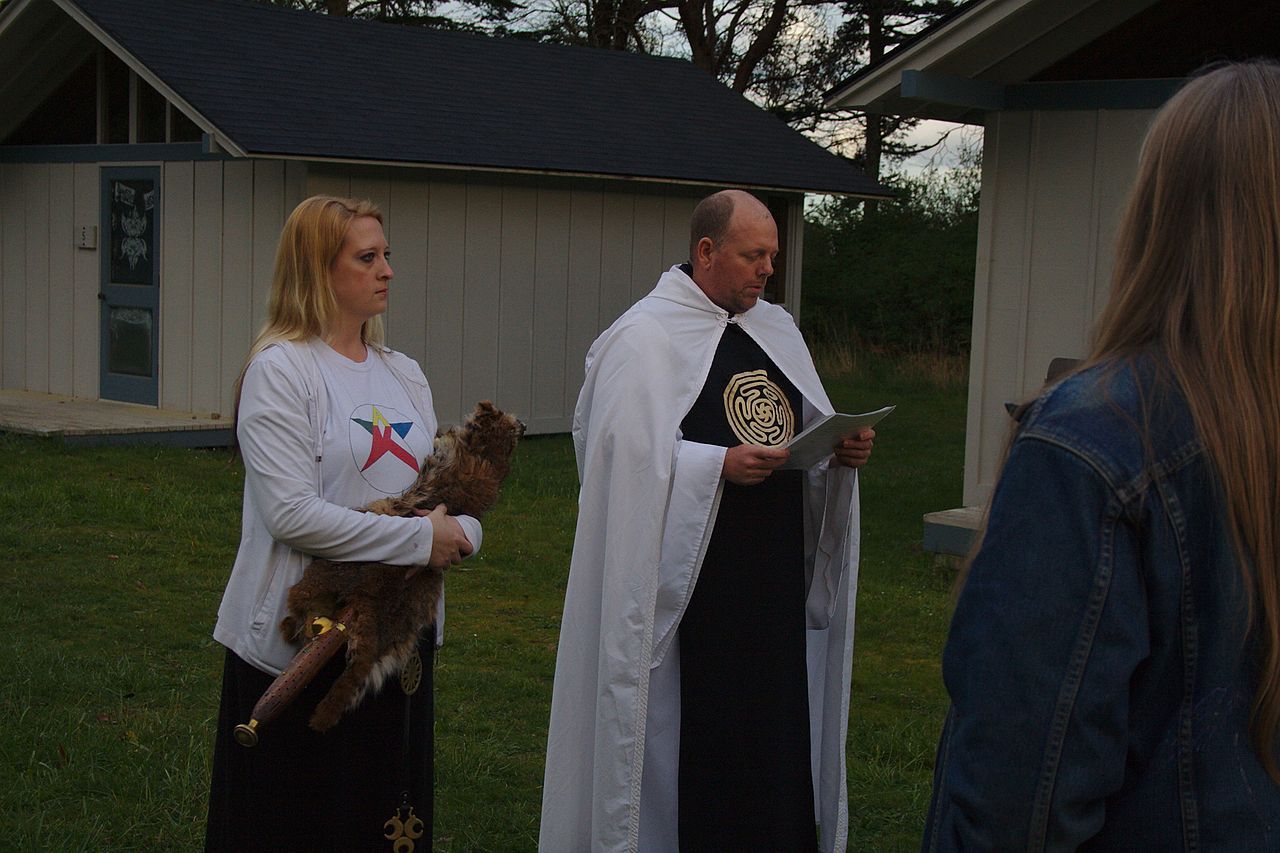 A man in a white robe is standing next to a woman, running a service for the Aquarian Tabernacle Church.