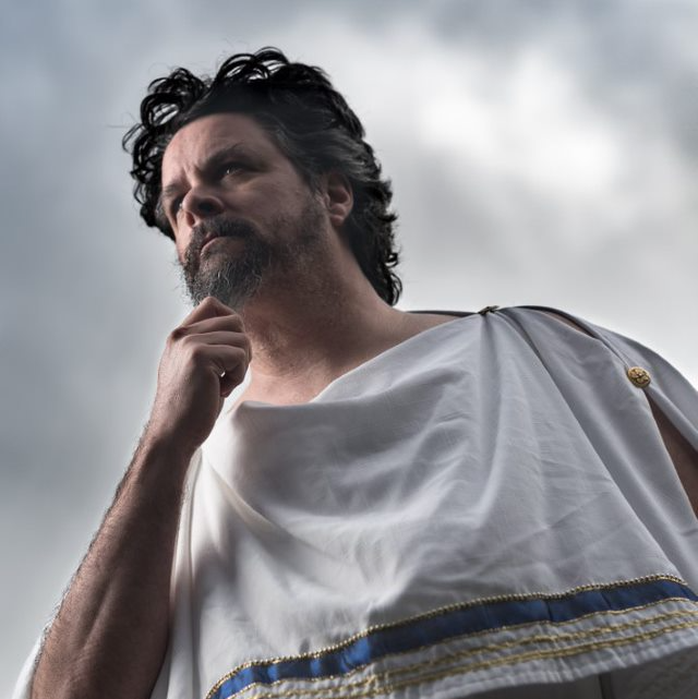 A man with a beard is wearing a white toga, representing Zeus.