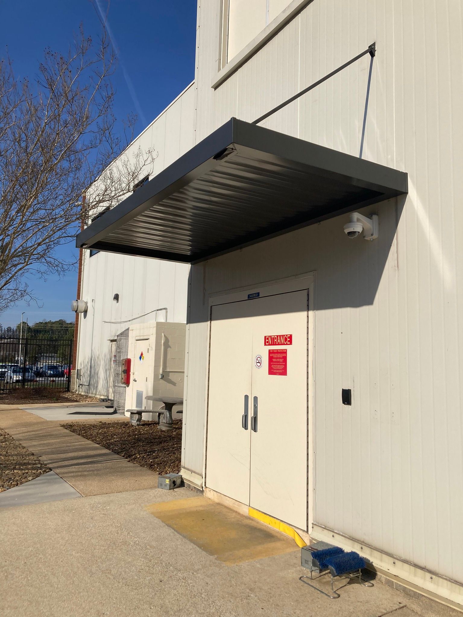Exterior view of a white building with a black awning over double doors, concrete pathway and a blue mat.