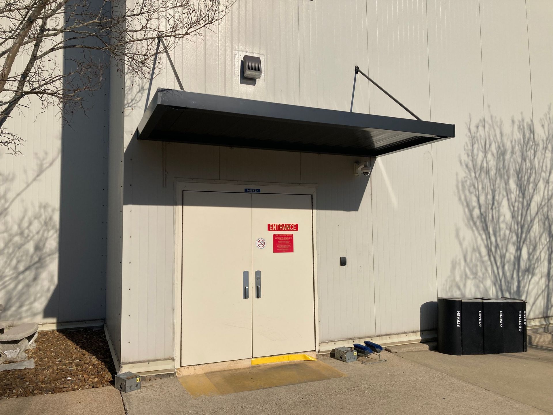 Exterior of a building with double doors, covered by a black awning. Black bins sit to the right.