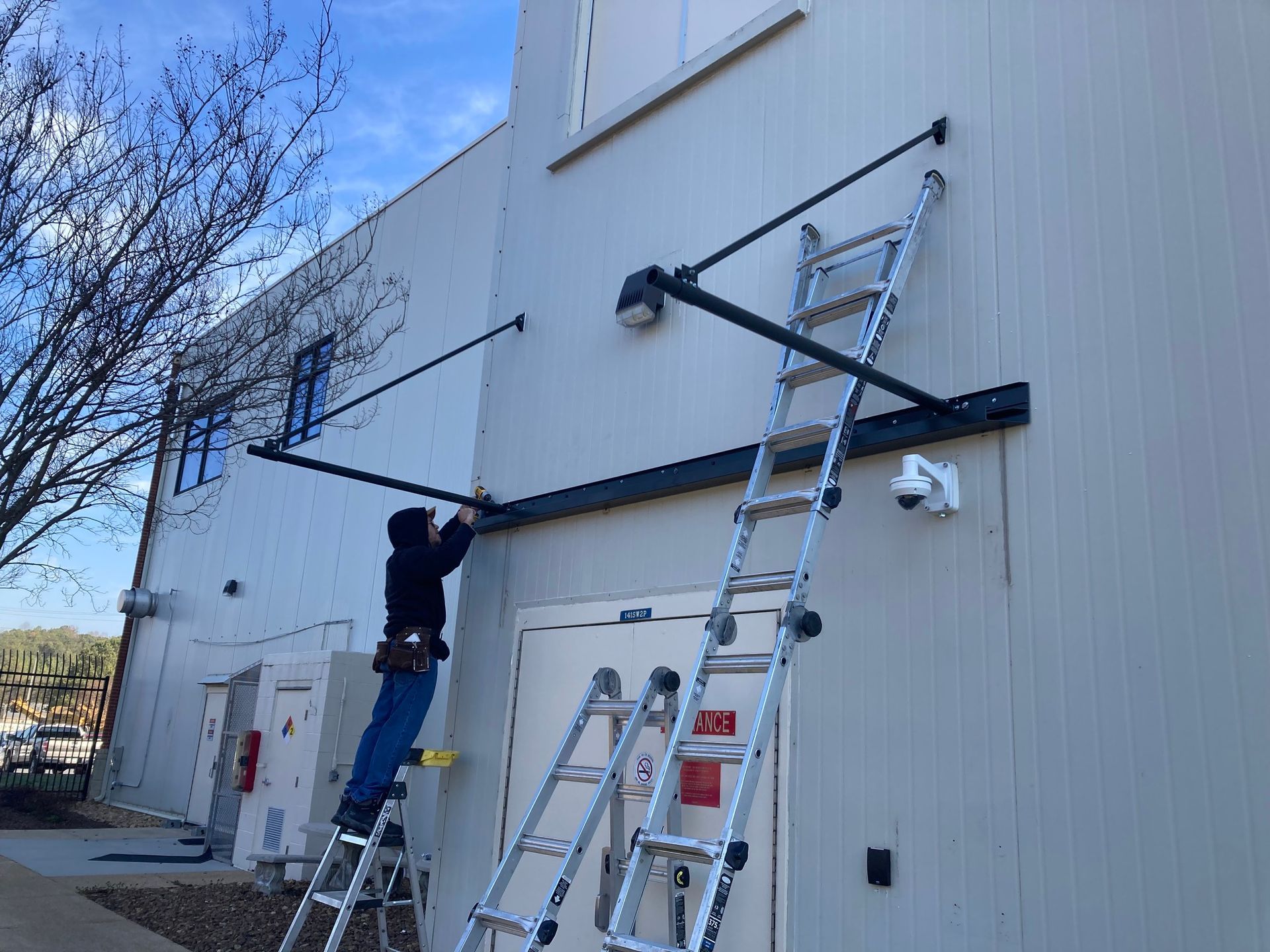 Person on ladder installing awning on a building. Gray siding, black awning frame, clear sky.