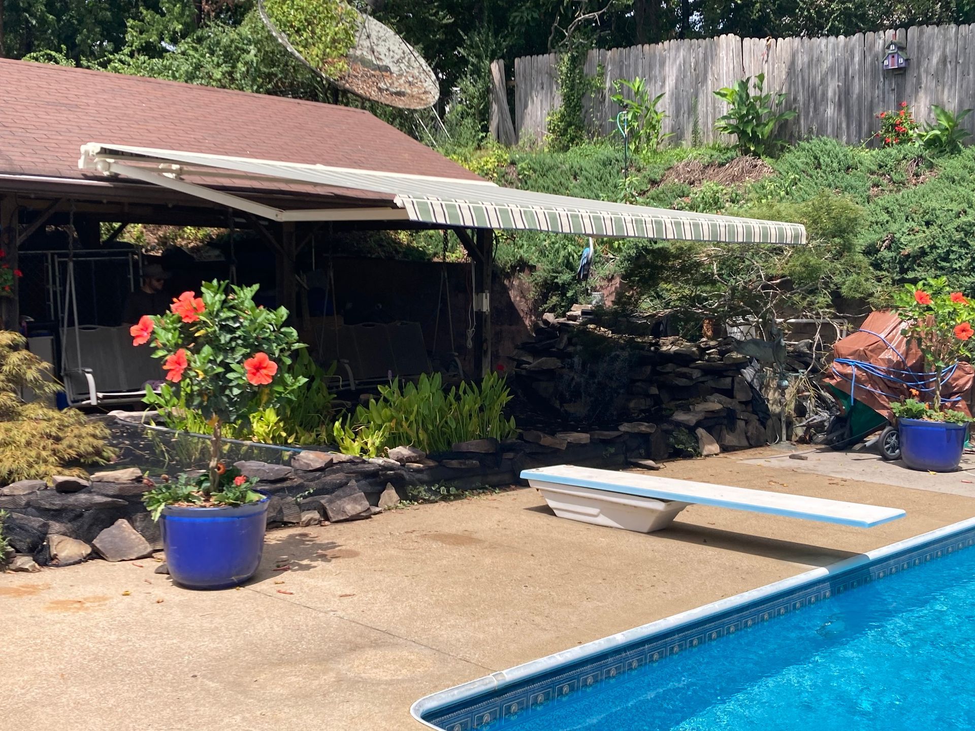 Backyard pool scene with a diving board, awning over a shaded area, and colorful potted plants.