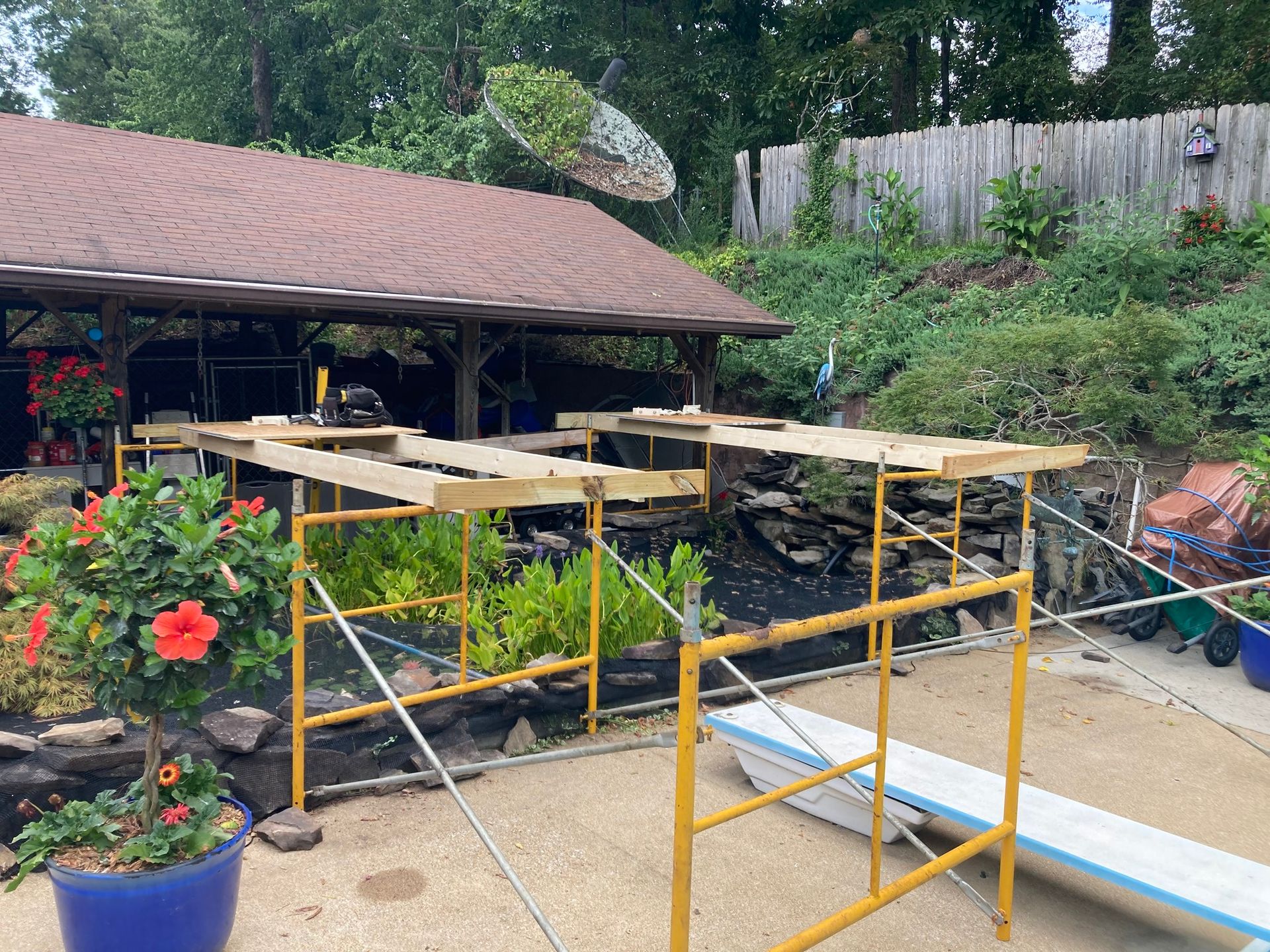 Yellow scaffolding supports wooden platforms near a brown-roofed structure with blooming flowers and a water feature.