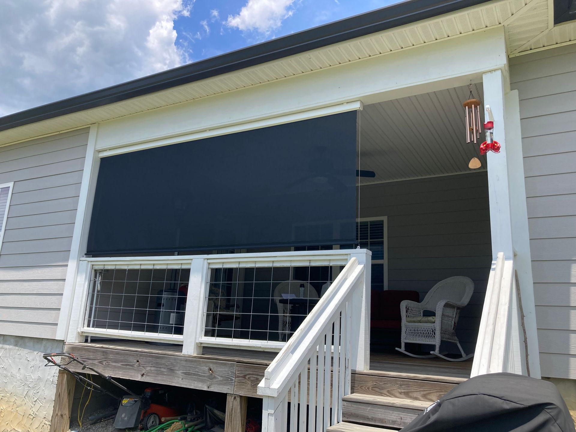 Black outdoor shade partially covers a porch. White railing and steps lead to seating and decorative wind chimes.