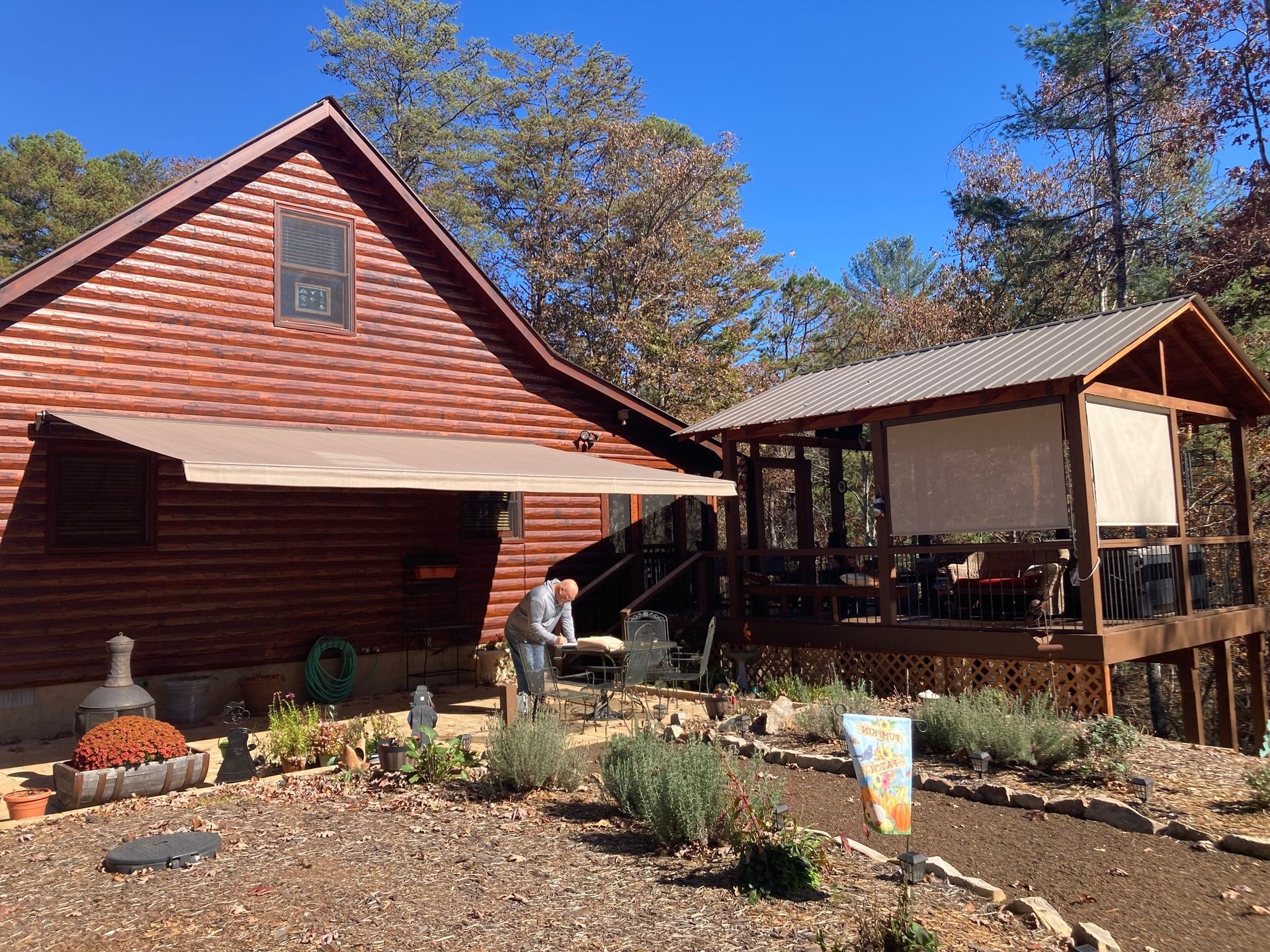 Log cabin with attached deck and awning; person on patio. Autumn foliage, blue sky.
