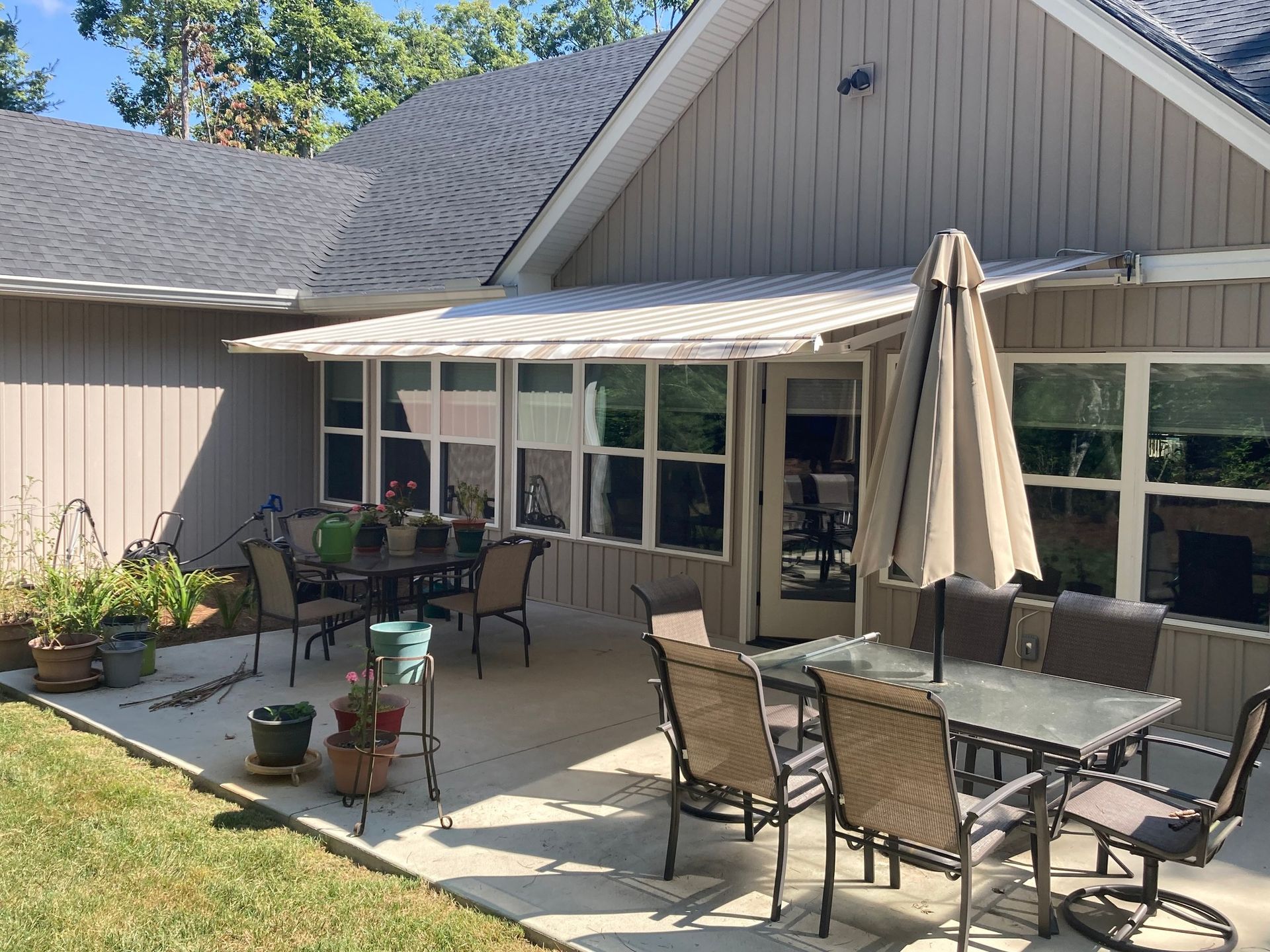 Outdoor patio with tables, chairs, and umbrella under a white awning. The patio is next to a house with large windows.