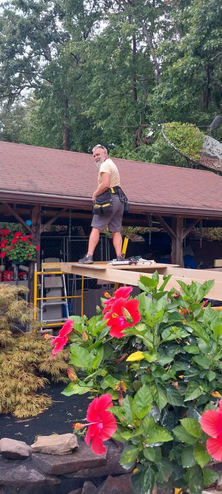 Man with tool belt on a roof, looking back. Red flowers in the foreground.