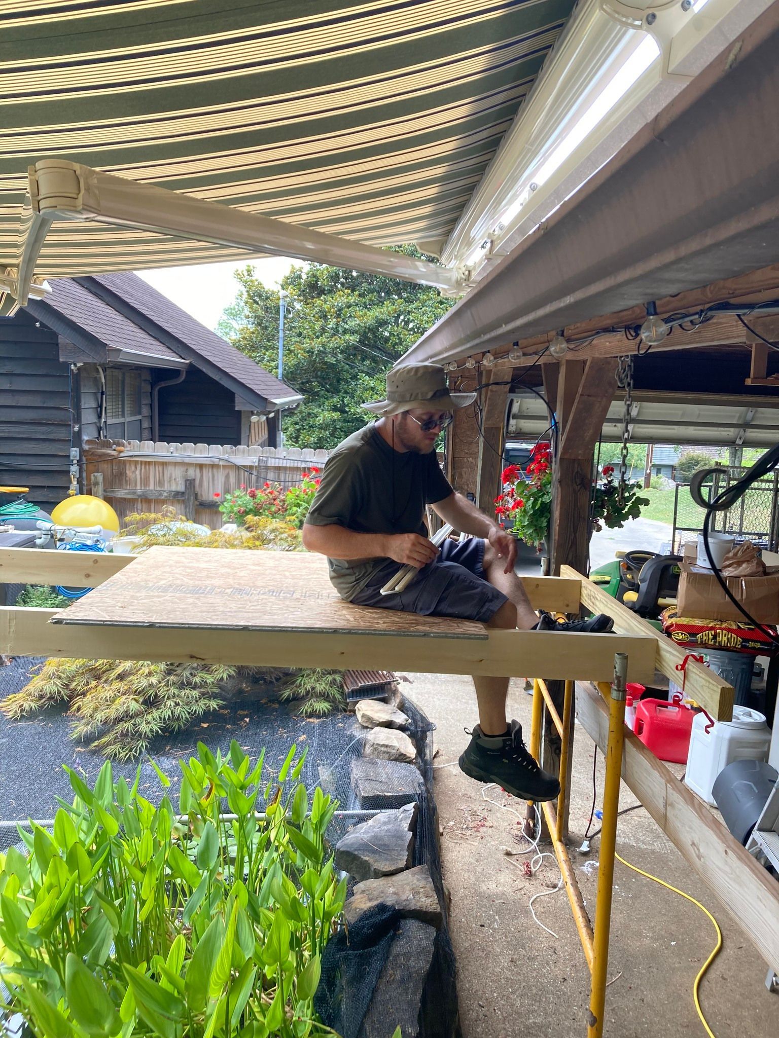 Man on scaffolding, working on a project near a garden. The setting is outdoors.