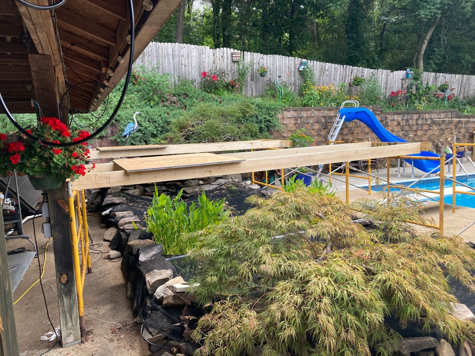 Deck construction over a rock wall and garden, near a pool with a slide.